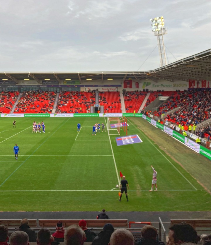 A Doncaster Rovers player gets ready to take a corner in front of the South Stand of the Eco Power Stadium during a match against Gillingham. Players from both teams; Rovers' wearing red and white hoops, Gillingham wearing blue, wait in the penalty area.