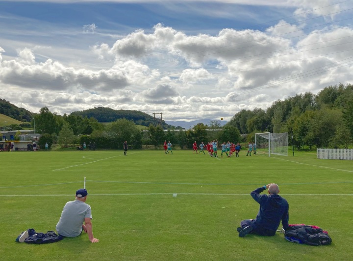 Two men sit on the grass to watch a football match between Mochdre Sports and Chirk AAA