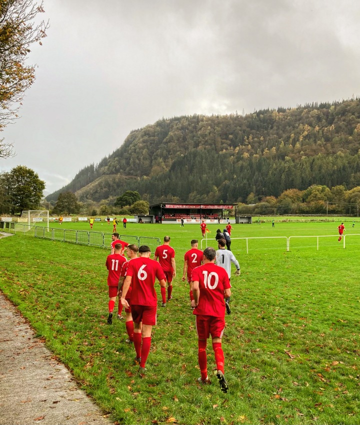 Llanrwst United's players head back out onto the field for the second half of their match. There are autumn trees on a hillside behind the pitch