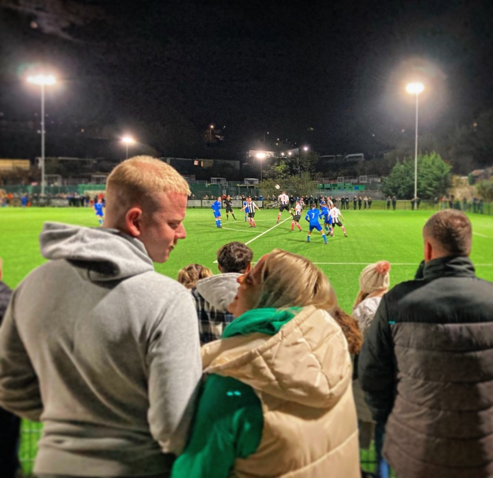 A young man and young woman look at each other on the terrace as Penydarren play a night match against Trethomas Bluebirds