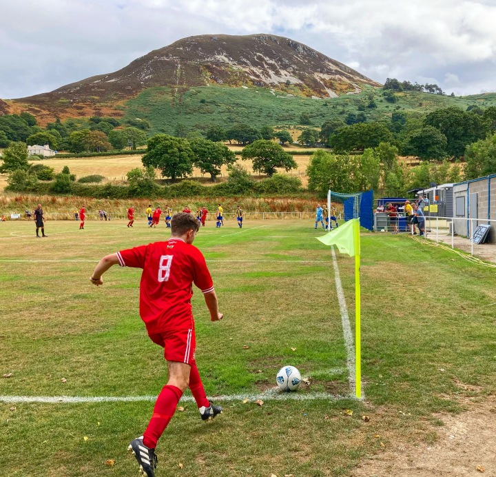 A Llanrug United player wearing number 8 takes a corner at Penmaenmawr's Cae Sling ground, with a large mountain in the background.