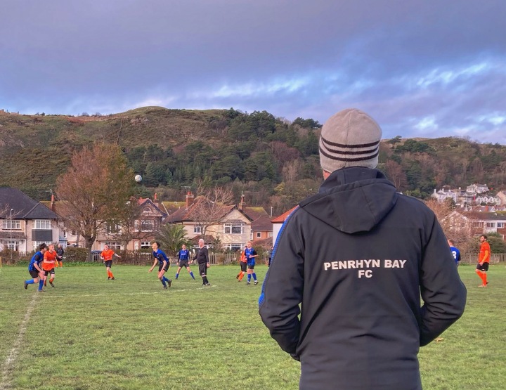 A man in a Penrhyn Bay FC hoodie watches his team play against Llysfaen