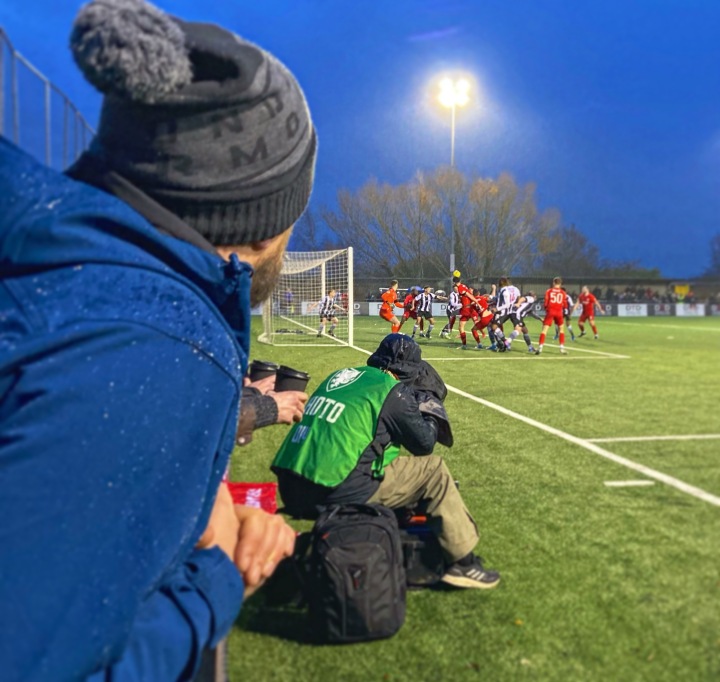 A supporter in a bobble hat and a photographer in waterproof clothing watch Flint Town United defend a Connah's Quay Nomads corner