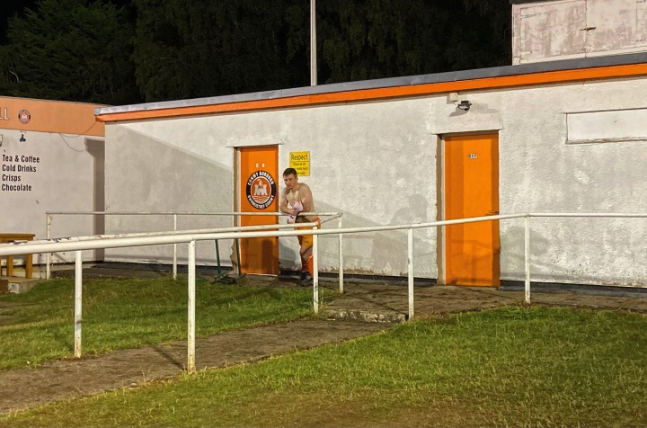 A shirtless substituted Conwy player watches the final minutes of Conwy v Colwyn Bay from in front of the changing rooms